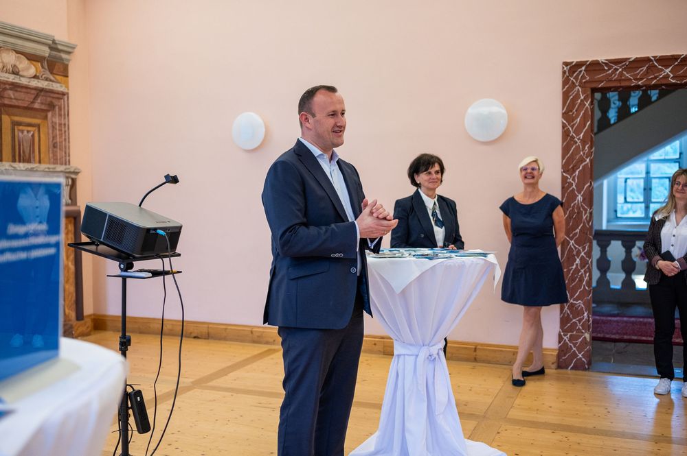 A man in a dark suit speaks with raised hands beside a draped standing table while three people look on in a bright reception hall.