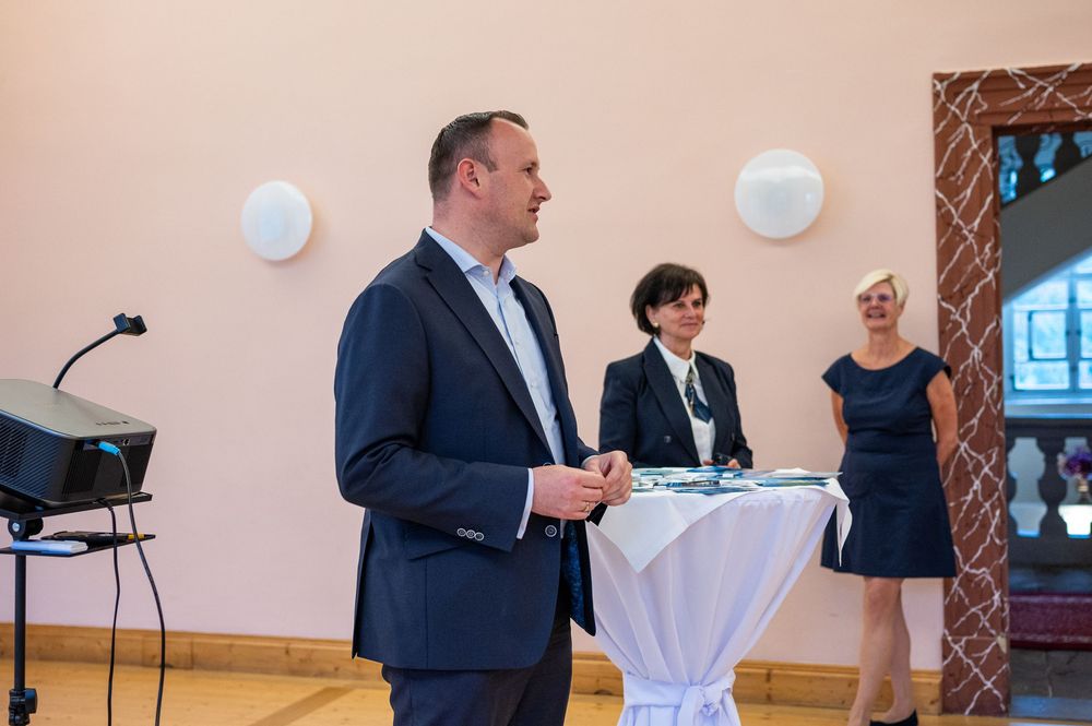 A man in a navy blazer stands speaking at an event beside a draped table while two women look on in a bright, elegant hall.