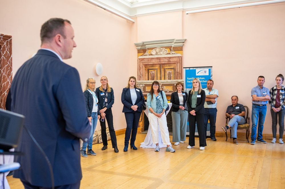 A man in a suit speaks to a group of people standing in a bright, ornate room with wooden floors during a gathering or event.