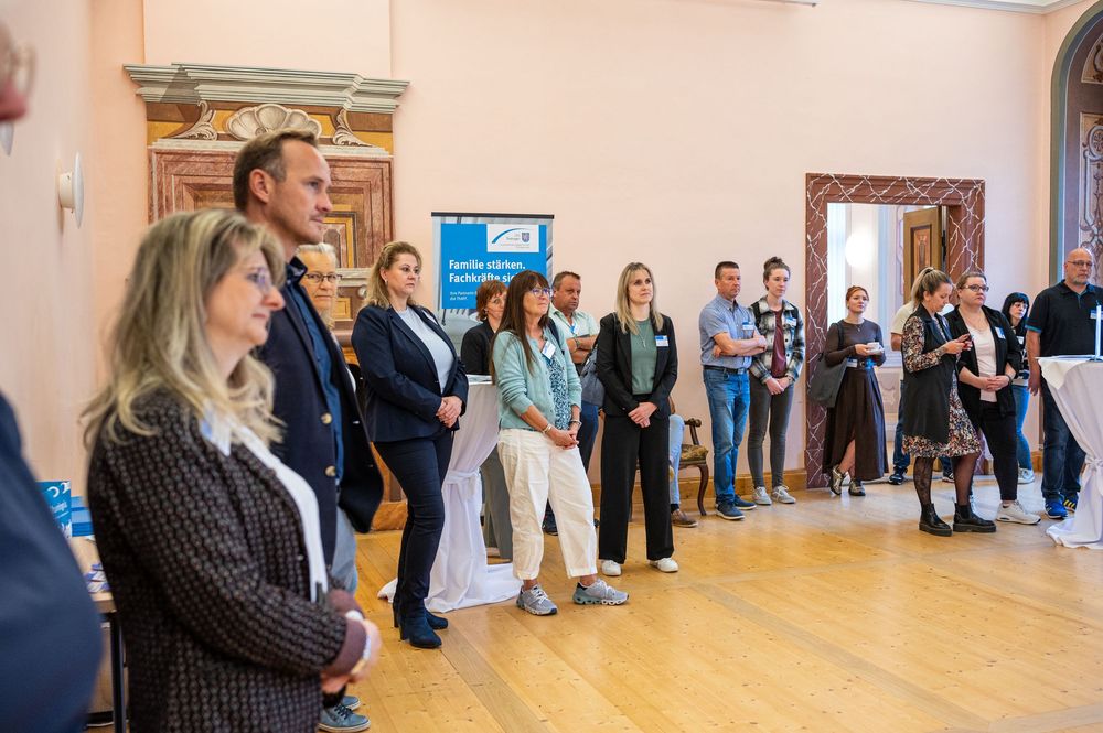 Group of business professionals standing and chatting in a bright hall with wooden floors and ornate walls during a networking event.