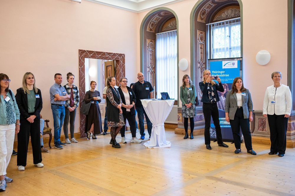 A group of people stand and chat in an elegant hall with arched windows, a polished wooden floor, and a high white cocktail table.
