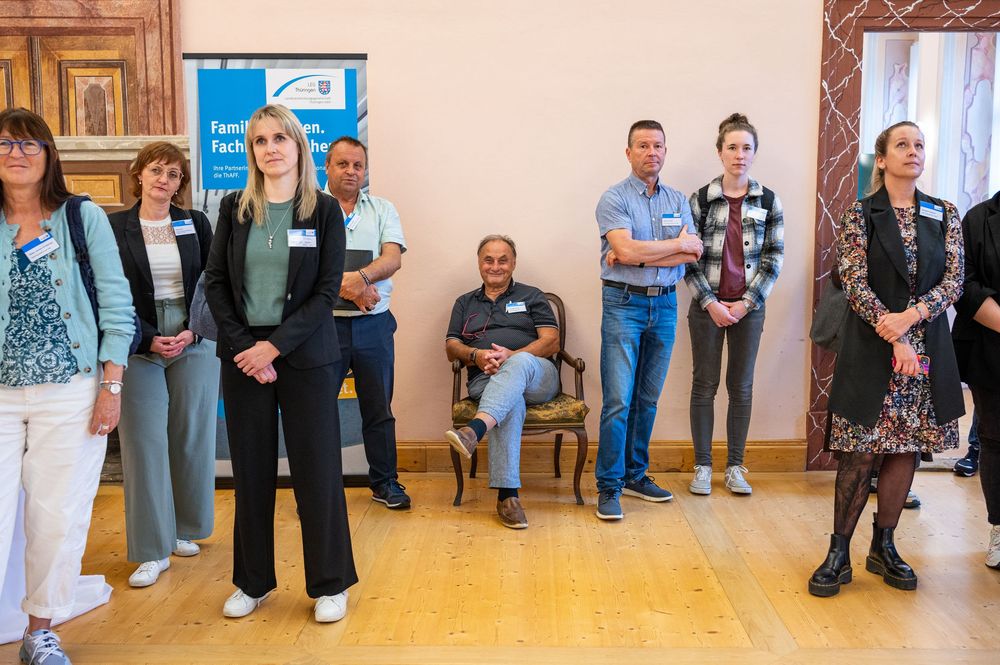 Group of attendees wearing name tags stand and sit in a wood-paneled room with parquet flooring during an indoor event or reception.