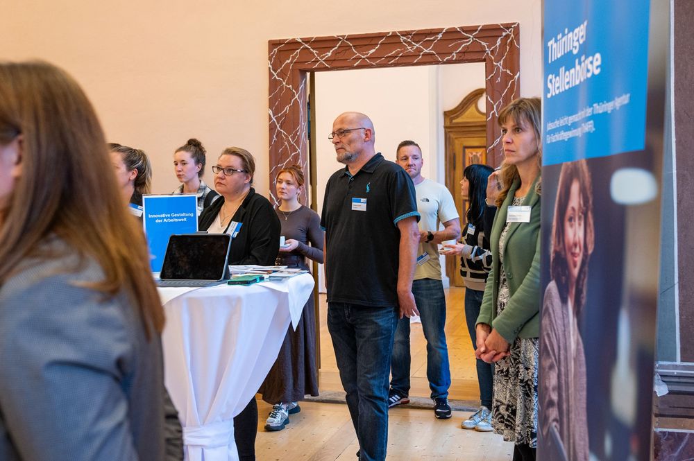 Attendees stand and chat around a white-draped table with a laptop at an indoor networking event, with a blue banner nearby.
