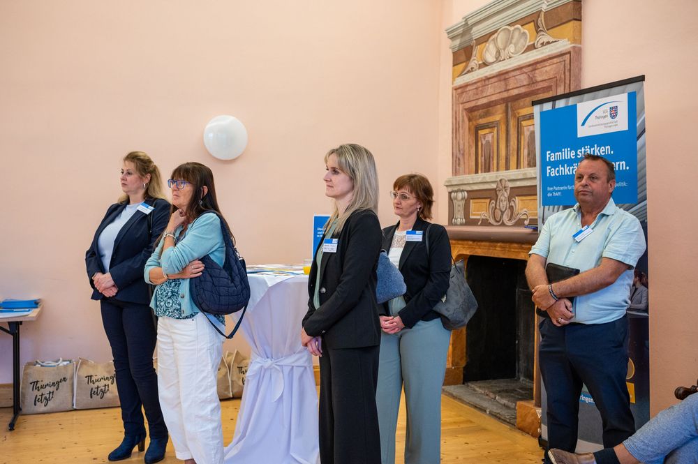 Five professionals in business attire stand in a bright room beside a draped table and a blue event banner during a networking gathering.