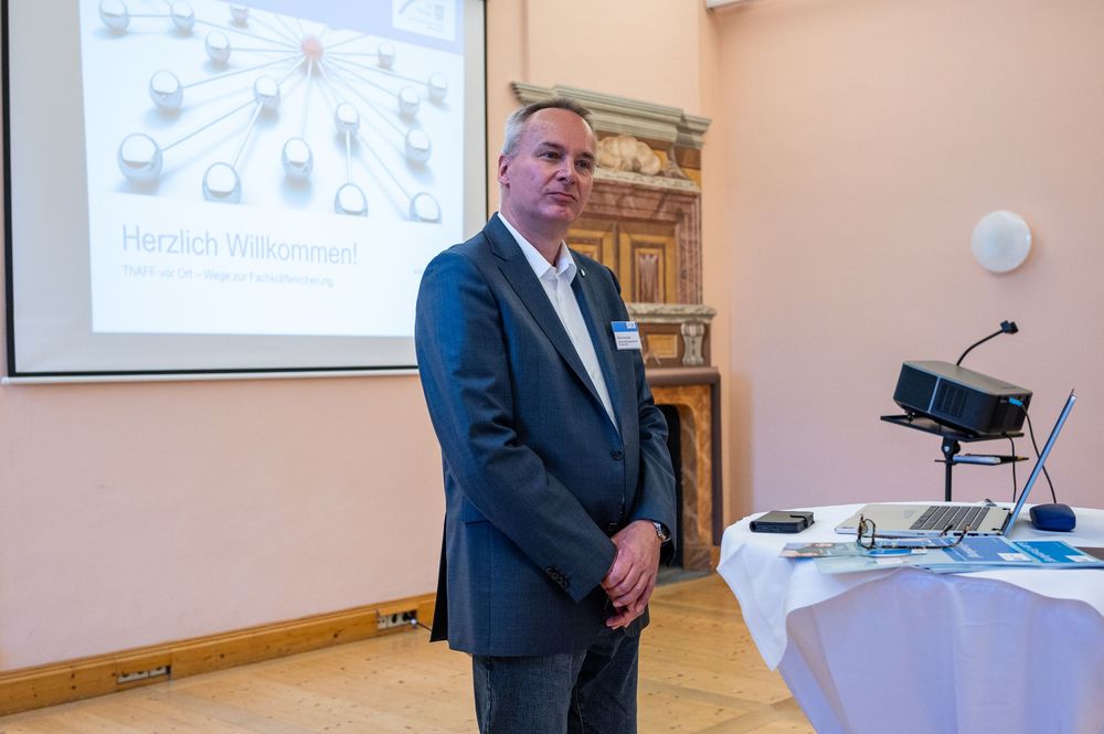 Man in a gray blazer stands beside a projector screen displaying a German welcome slide during a business presentation.