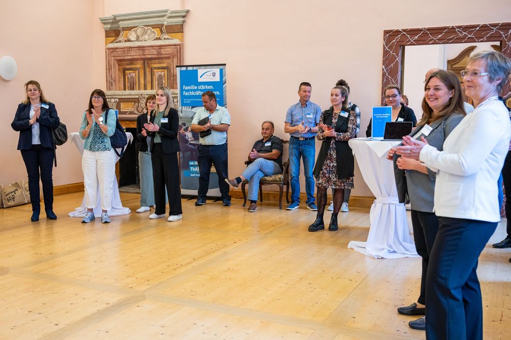 Guests stand and chat in a bright hall with a wooden floor during an event, with a woman in white speaking beside a draped table.