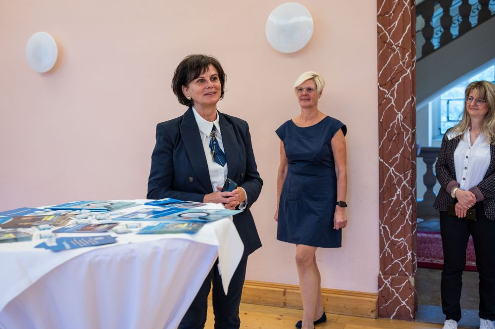Three women stand in a bright room with pink walls and round wall lamps, near a table with brochures, as one in a dark blazer smiles.