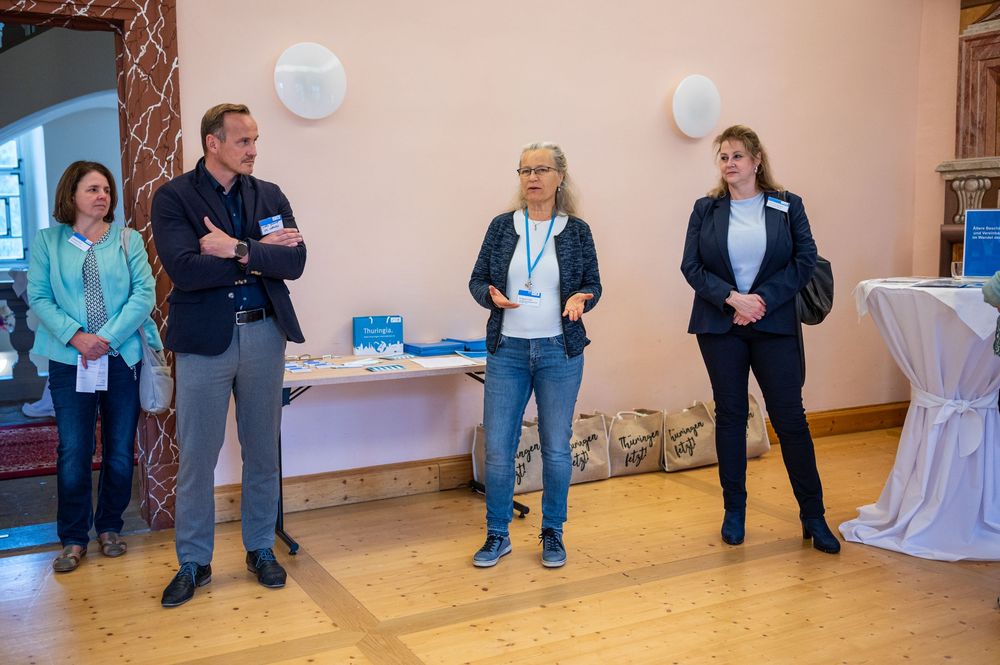 Four people wearing name tags stand in a bright event room with wooden floors, listening to a speaker beside a table of materials.