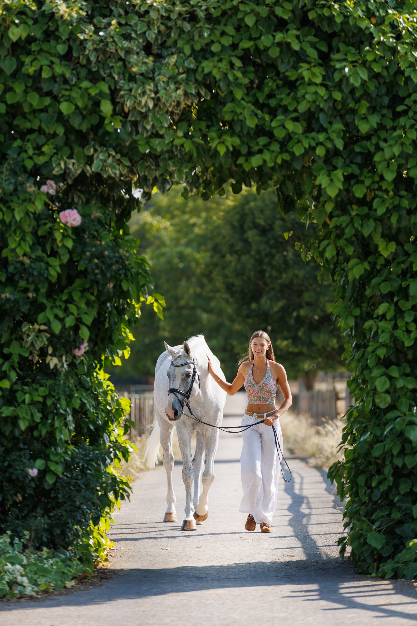 Woman in white lace dress walks alongside a brown horse on a sunlit forest path.