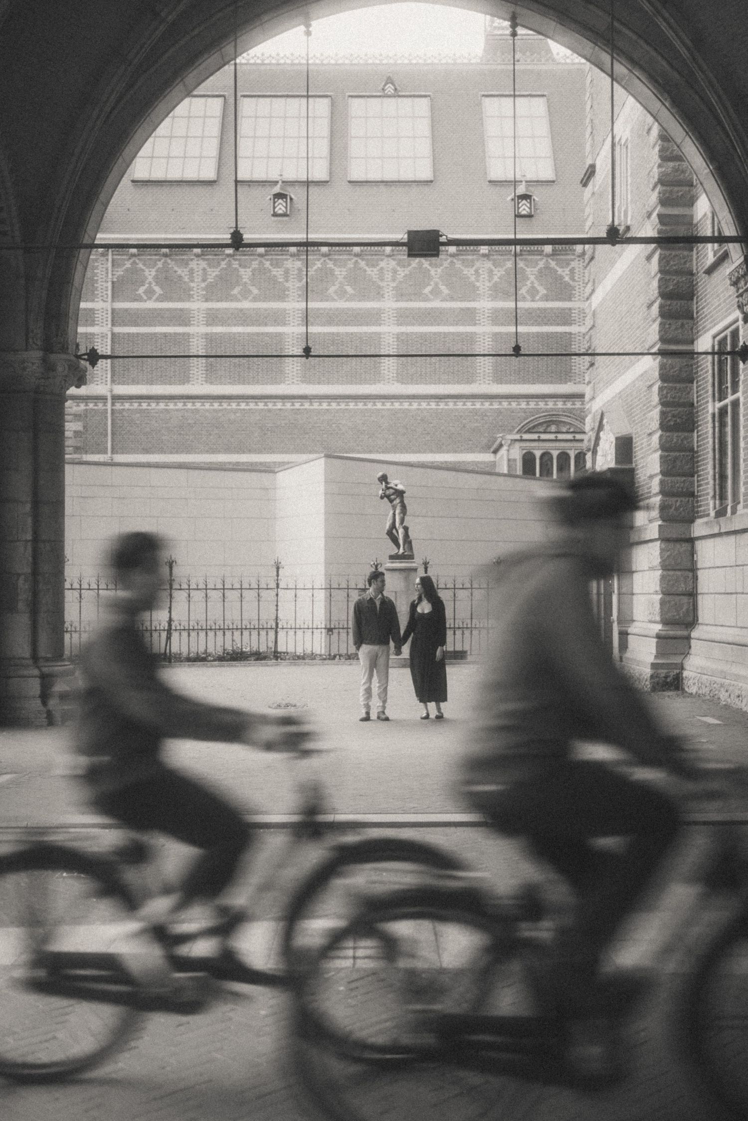 Two blurred cyclists ride through a stone archway while a couple holds hands near a statue in a sunlit courtyard. Rijksmuseum