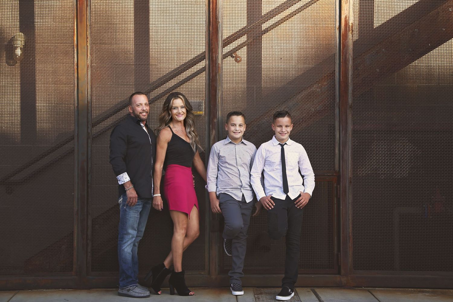 Family of four posing against a rustic rusted metal wall, with parents and two boys in dressy casual outfits.