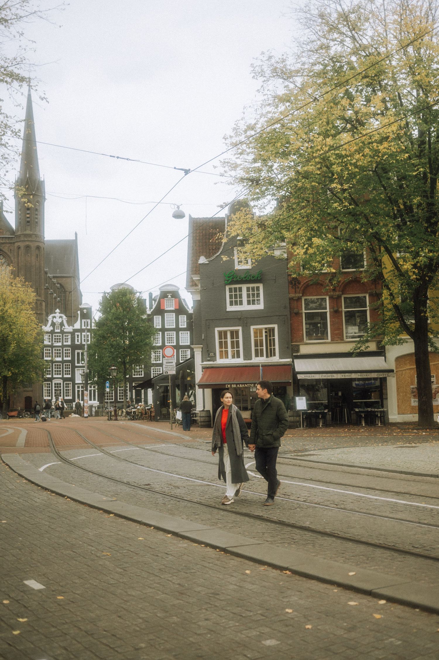 Couple walking hand in hand across a cobblestone square in Amsterdam with a Gothic church and canal houses behind them on an autumn day.
