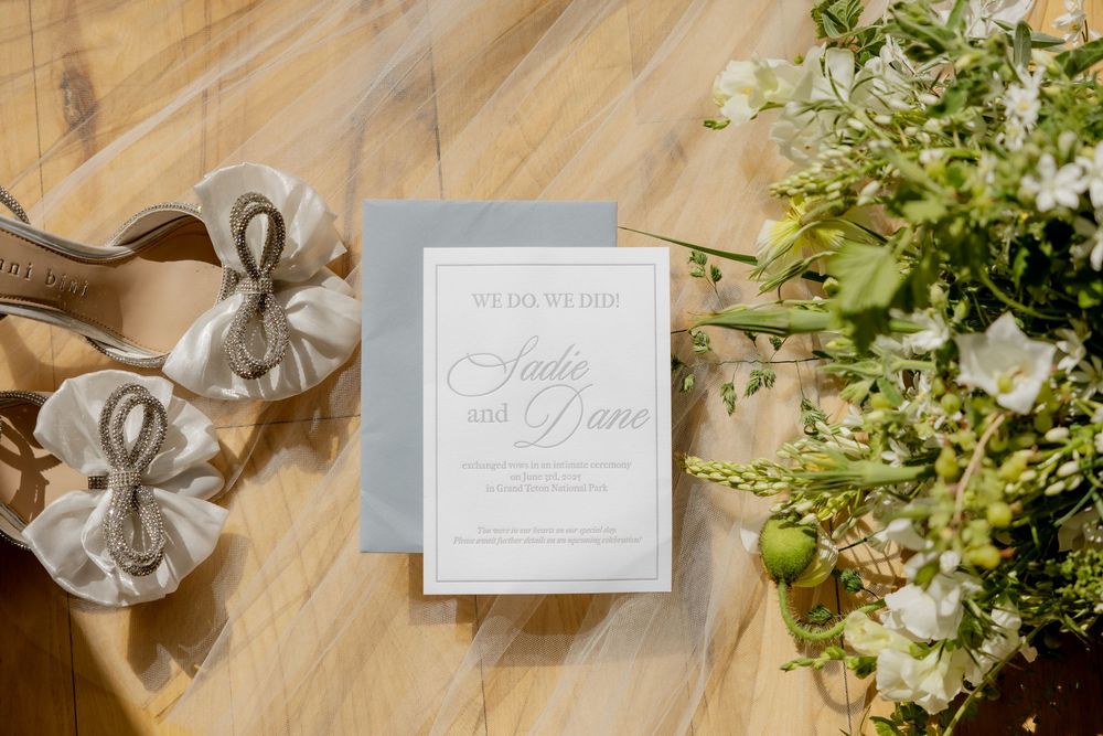 Wedding invitation for Sadie and Dane on a gray card atop a wood floor beside silver bow sandals and a white and green floral bouquet.