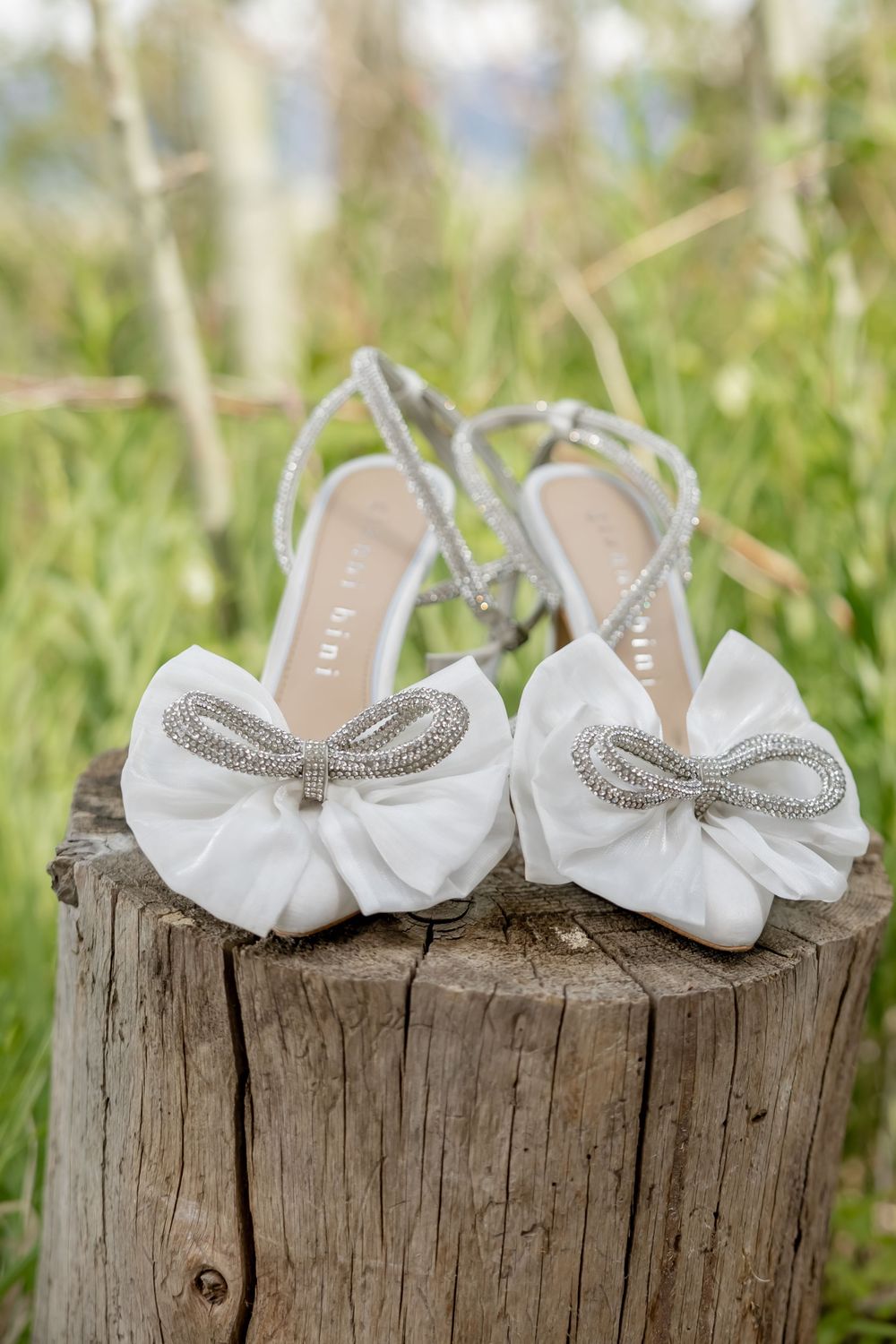 White bridal sandals with satin bow accents and rhinestone embellishments displayed on a rustic wooden stump in a green meadow.
