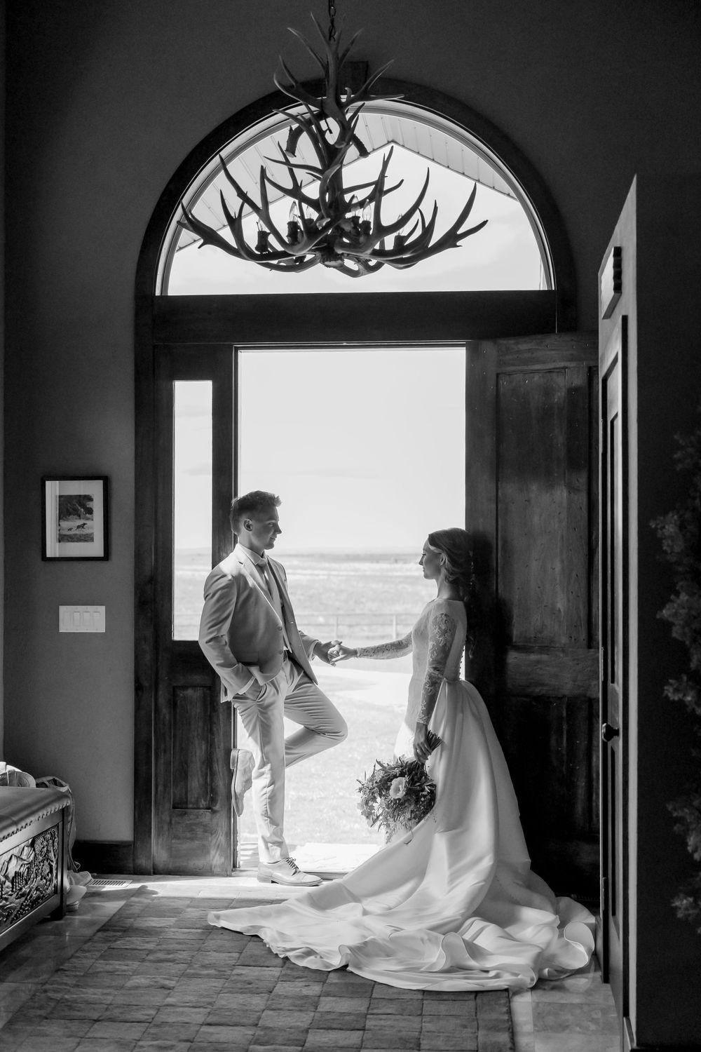 Black and white photo of a bride and groom holding hands in an arched doorway with an antler chandelier and ocean view beyond.