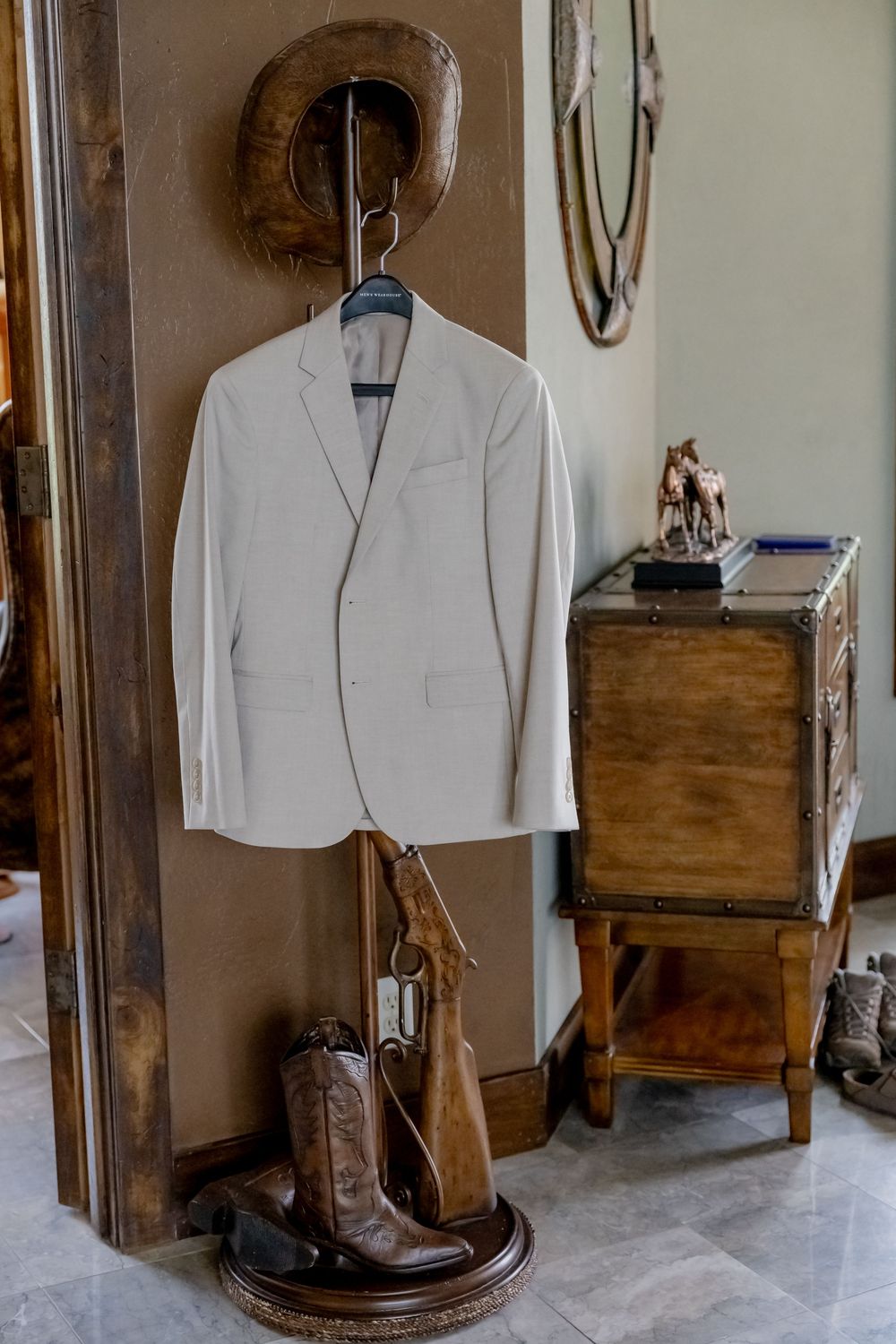 Light beige suit jacket hanging on a rustic stand with a brown cowboy hat and leather western boots beside a wooden chest.