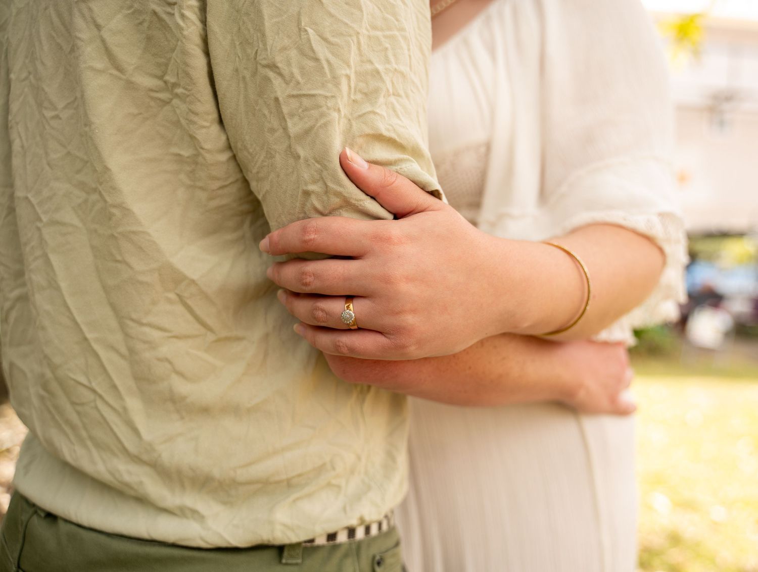 Pregnant woman in white dress embracing man in beige shirt, showing gold ring and bracelet.
