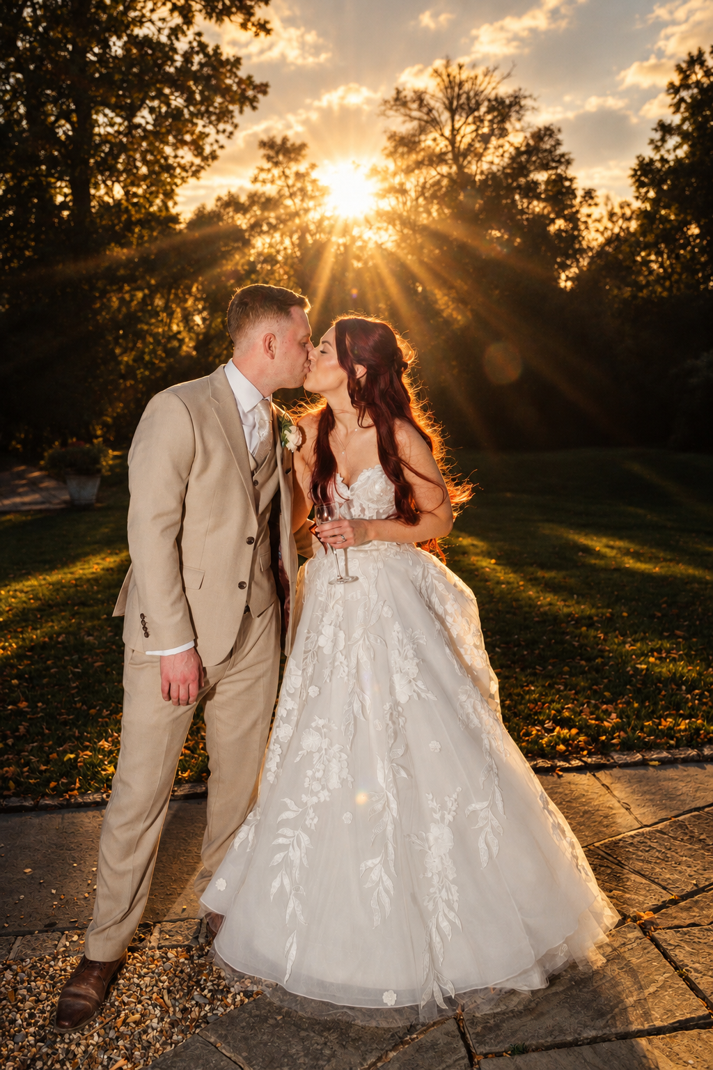 A bride and groom share a romantic kiss at golden hour sunset, surrounded by lush green trees in an outdoor garden setting.