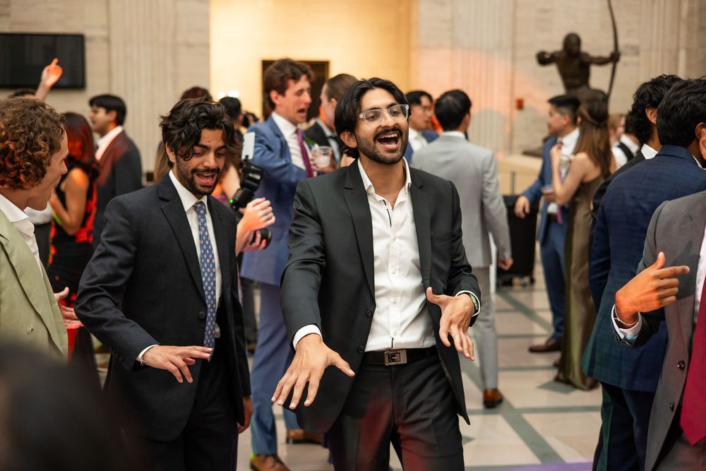 Two men in suits laugh and gesture at an elegant indoor social event with guests mingling in background.