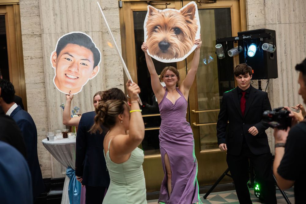 Women in formal dresses hold oversized face and dog cutouts at a lively wedding reception dance floor.