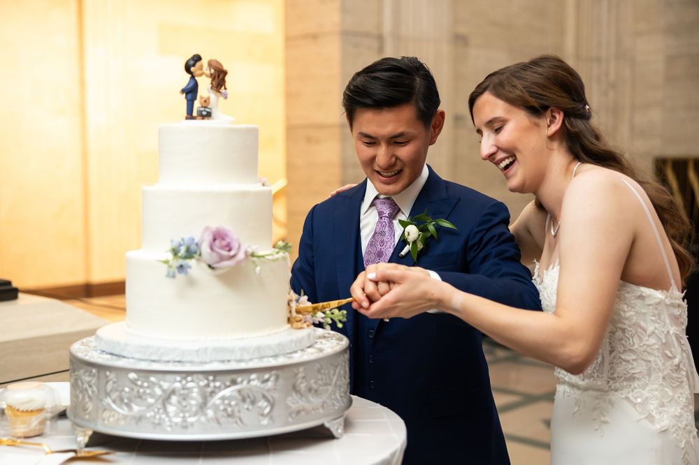Bride and groom smiling while cutting their white three-tier wedding cake topped with custom figurines at their reception.
