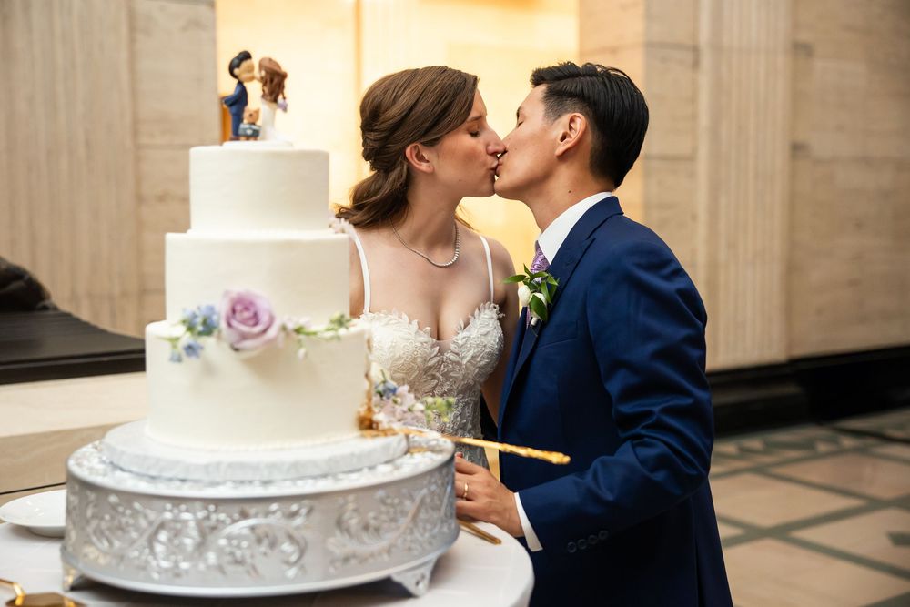Bride and groom share a kiss while cutting their elegant white wedding cake adorned with purple flowers.