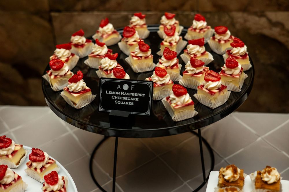 Lemon raspberry cheesecake squares displayed on a tiered black stand, topped with whipped cream and fresh raspberries.