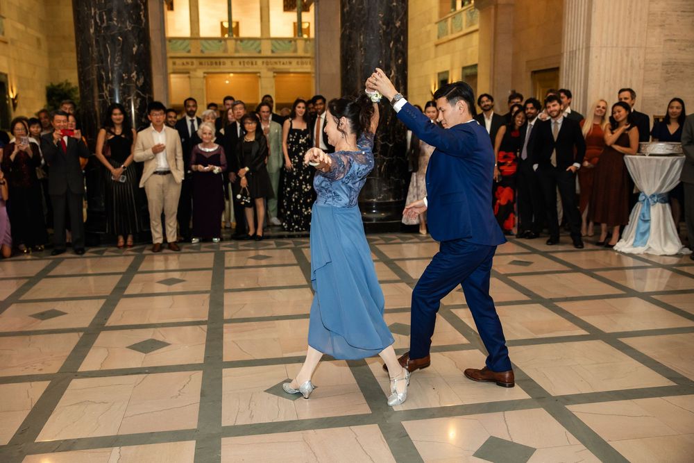 Couple dancing elegantly at a formal event in a grand marble hall with guests watching in evening attire.