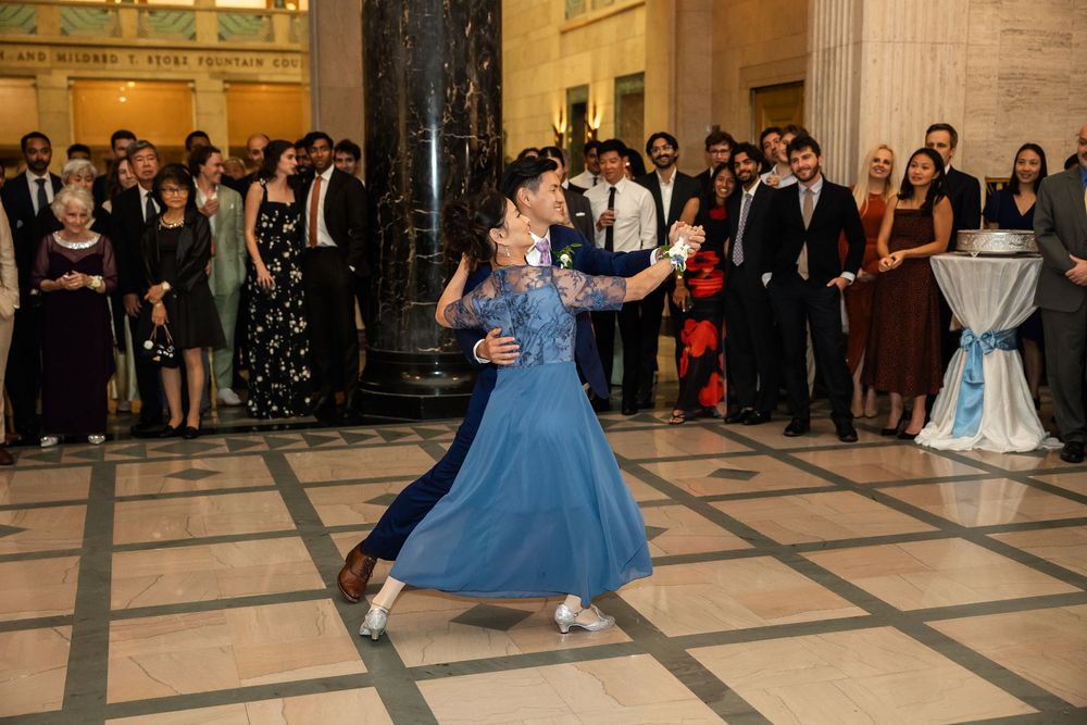 Elegant couple performing a ballroom dance in a grand hall while formally dressed guests watch in the background.