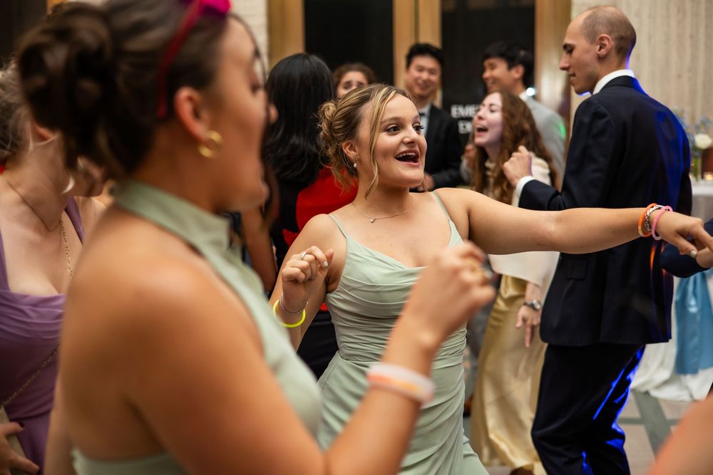 Teens in formal attire dance joyfully at a prom event, with a girl in a mint green dress smiling.