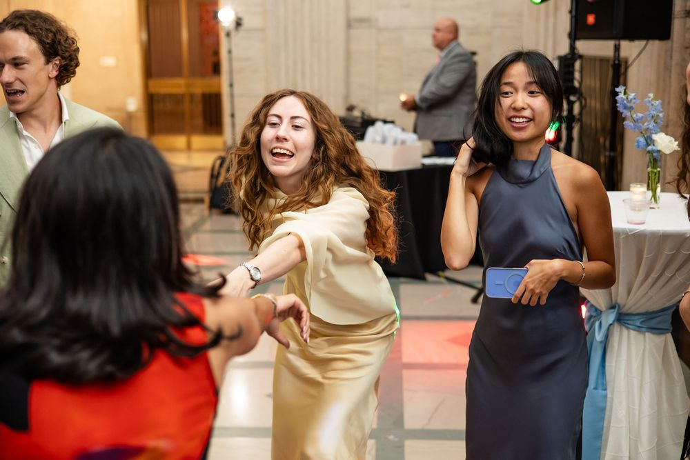 Women dancing and laughing at an elegant indoor party event with decorative flowers and guests in background.