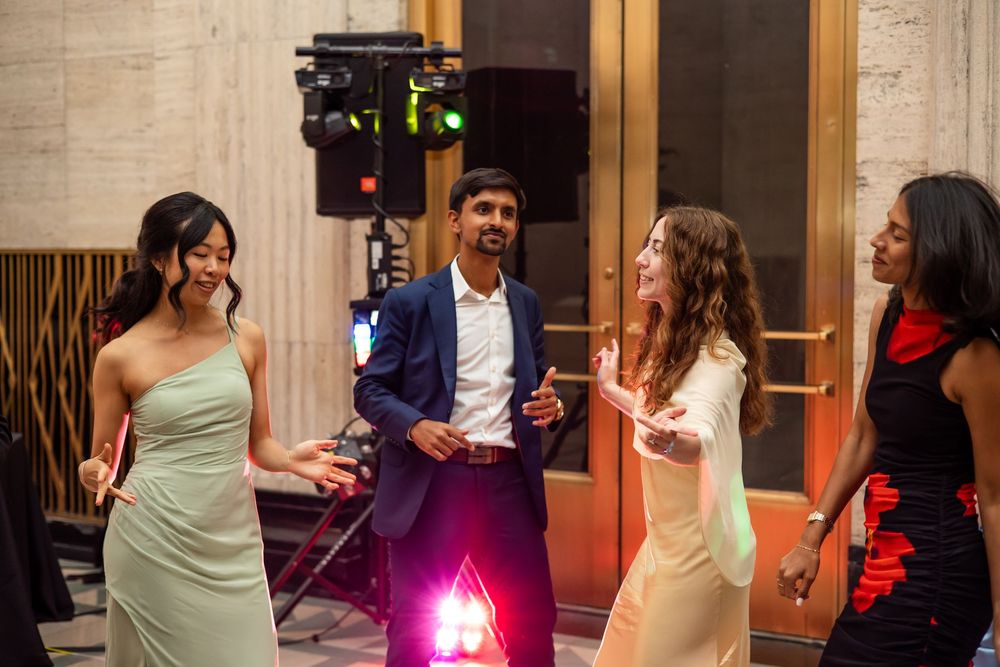 Four young adults in formal attire dancing and socializing at an elegant indoor event with colorful lights.