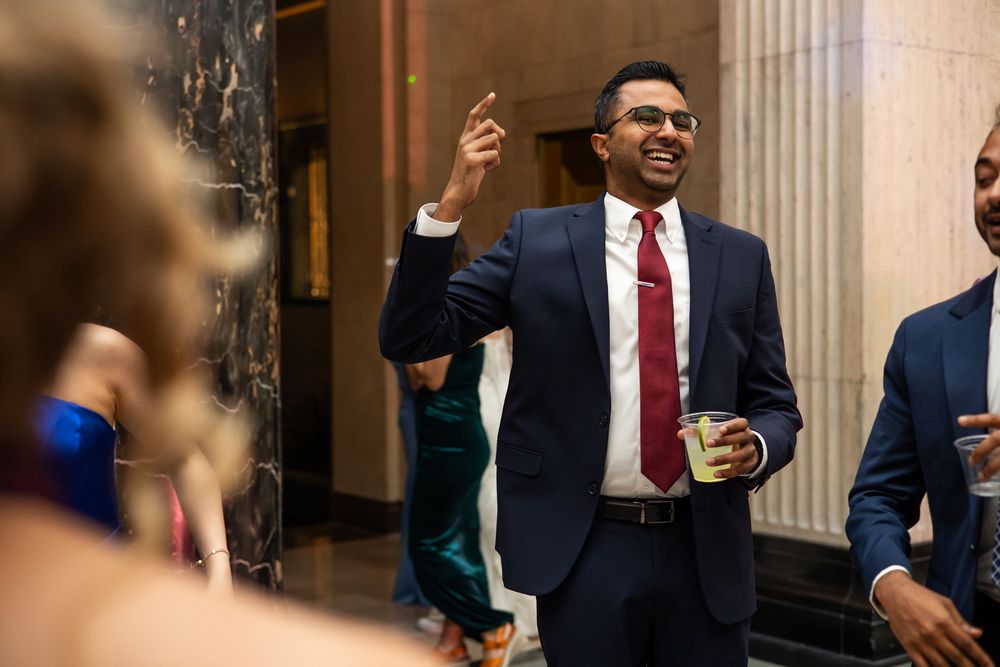 A man in a navy suit with red tie laughs and raises his hand at an elegant indoor event.