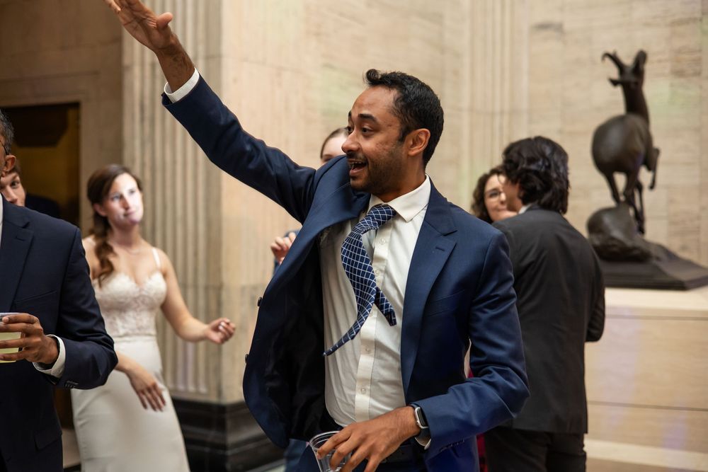 Man in blue suit waving at wedding reception inside elegant venue with columns.