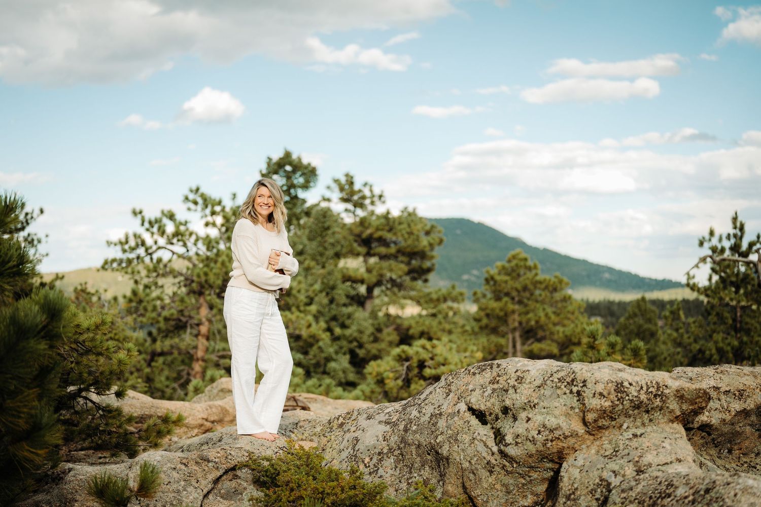 A woman in white stands on rocky terrain with pine trees and rolling hills under a partly cloudy sky.