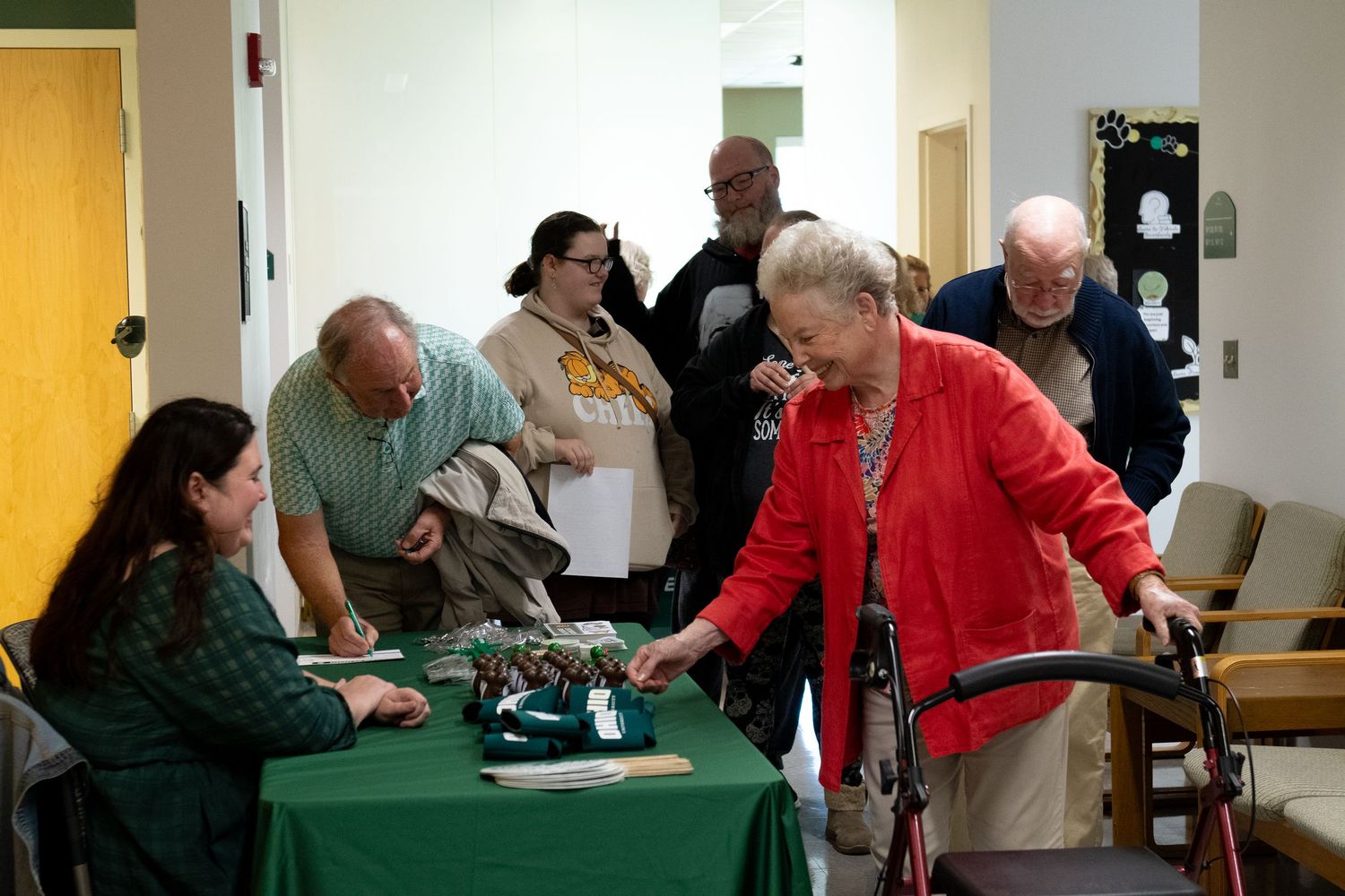 Elderly woman in red jacket at registration table with green tablecloth at indoor community event.