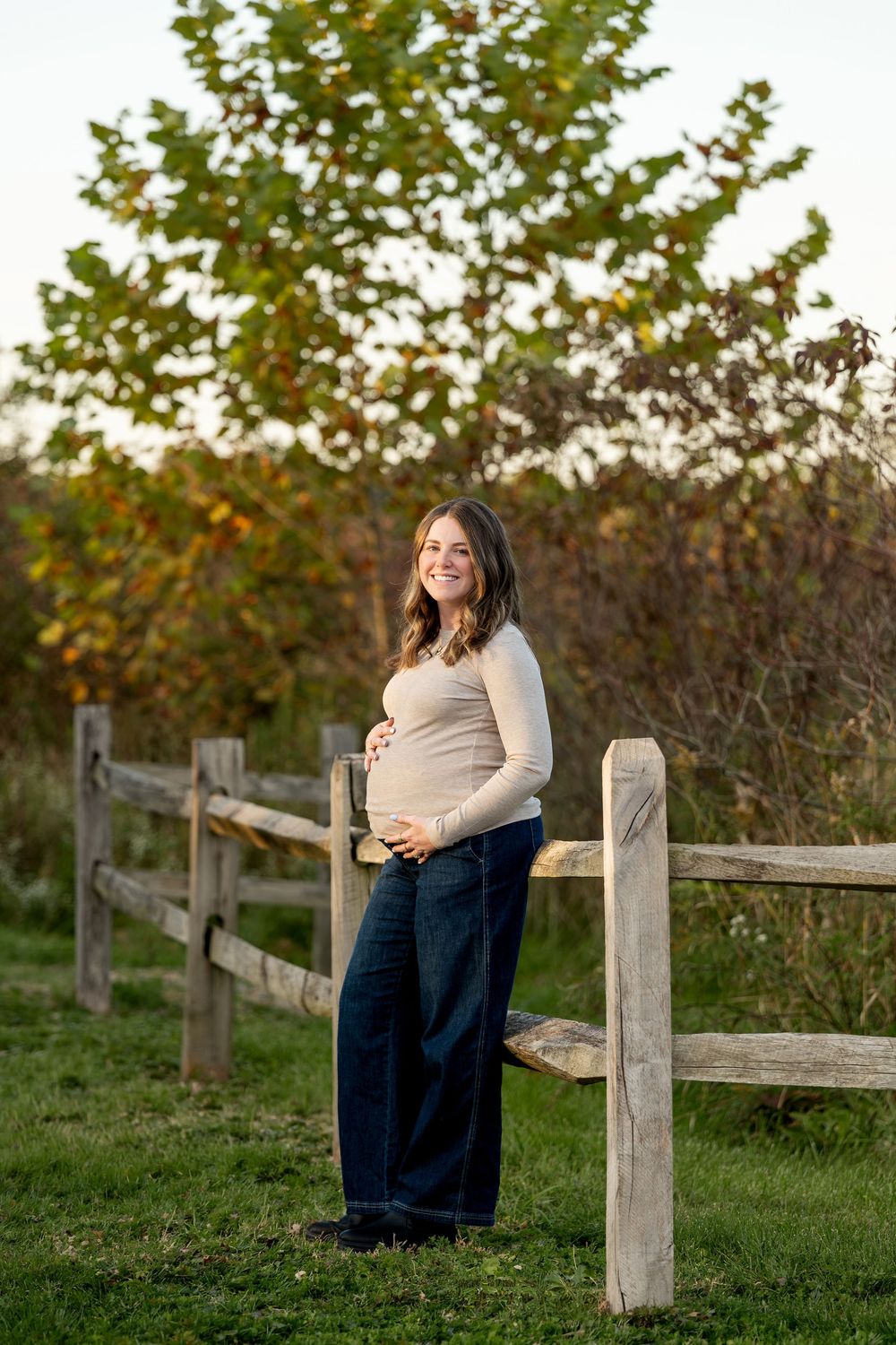 Smiling pregnant woman in beige top and jeans cradling baby bump by rustic wooden fence outdoors.