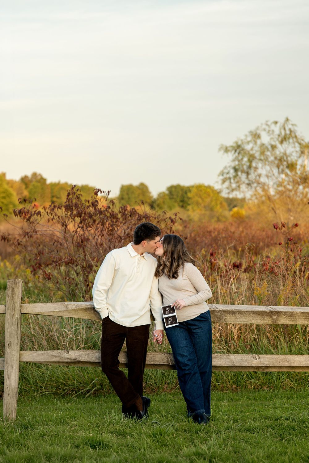 Couple kissing by rustic fence in autumn field, holding ultrasound photo for pregnancy announcement.