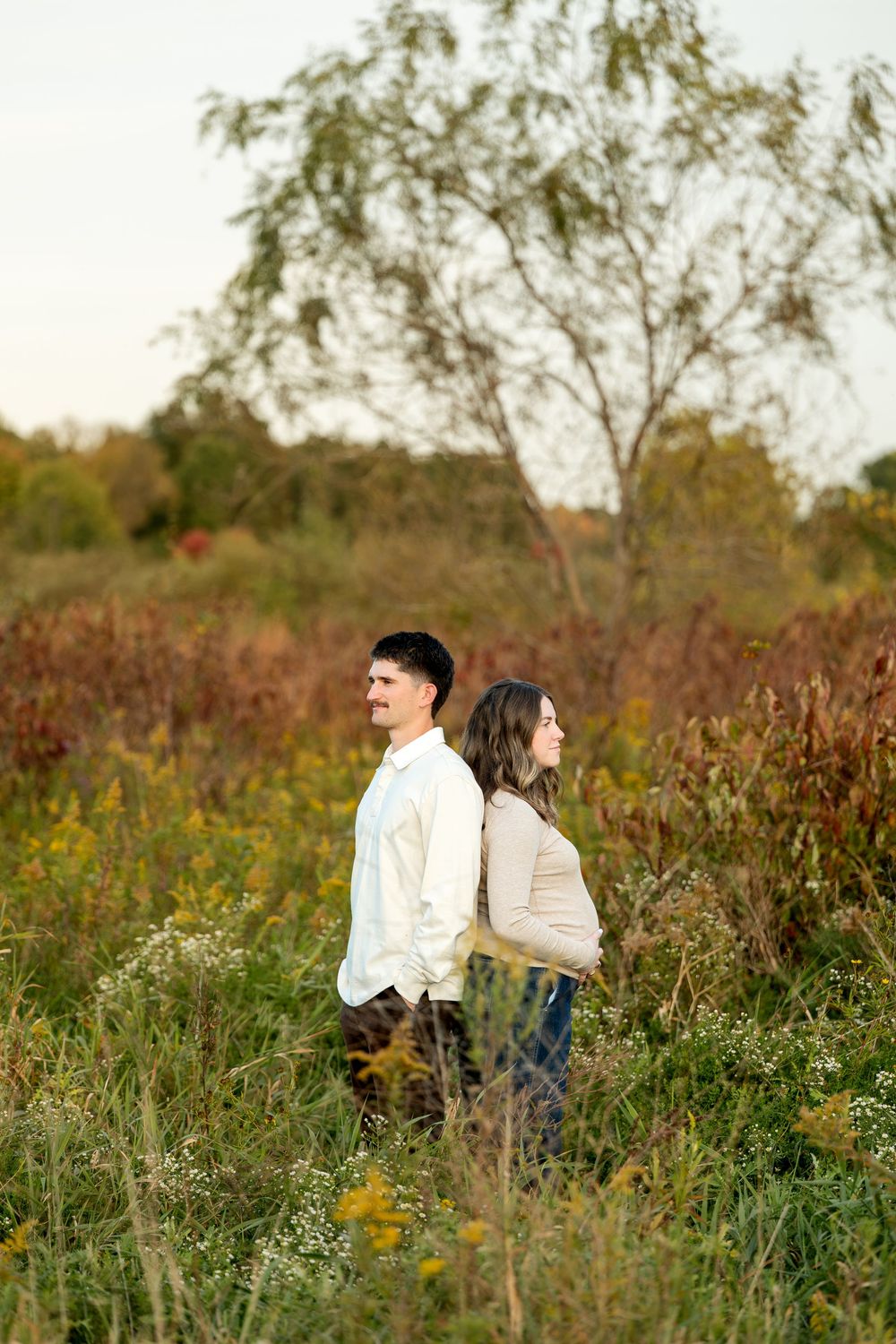 A couple stands back-to-back smiling in an autumn meadow with colorful foliage and a tree in the background.