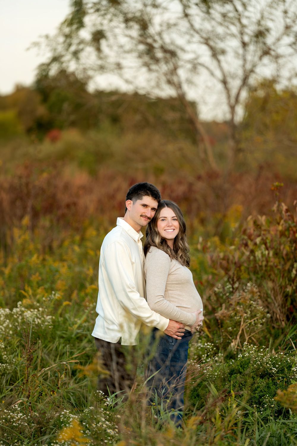 A couple poses in an autumn field, the woman visibly pregnant, both smiling warmly in neutral tones.