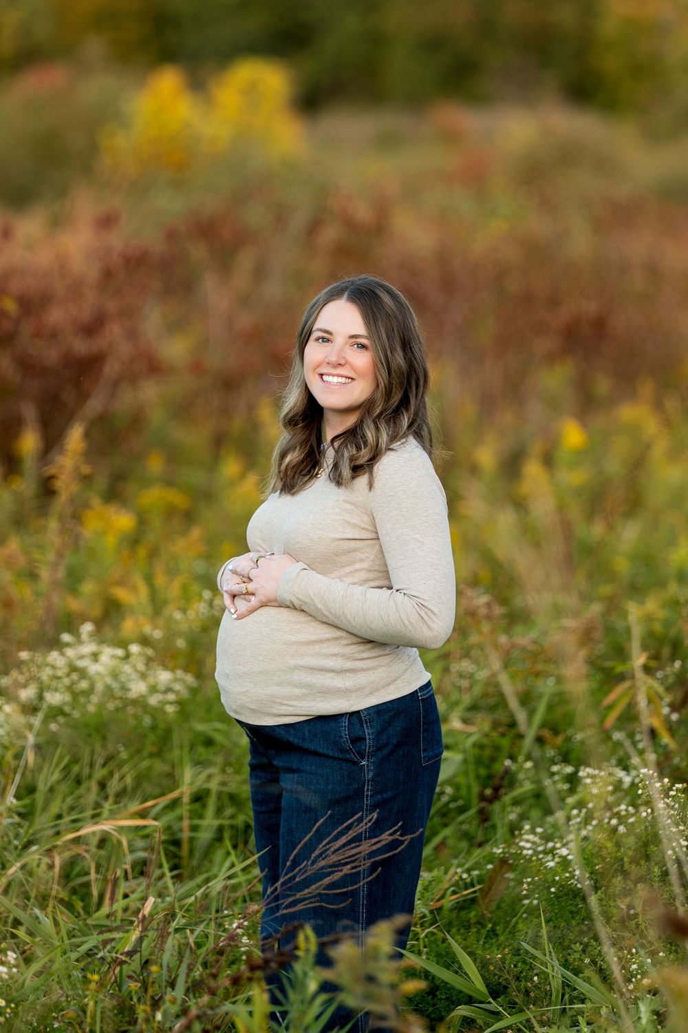 Smiling pregnant woman in a beige long-sleeve top holds her baby bump in a vibrant autumn meadow.