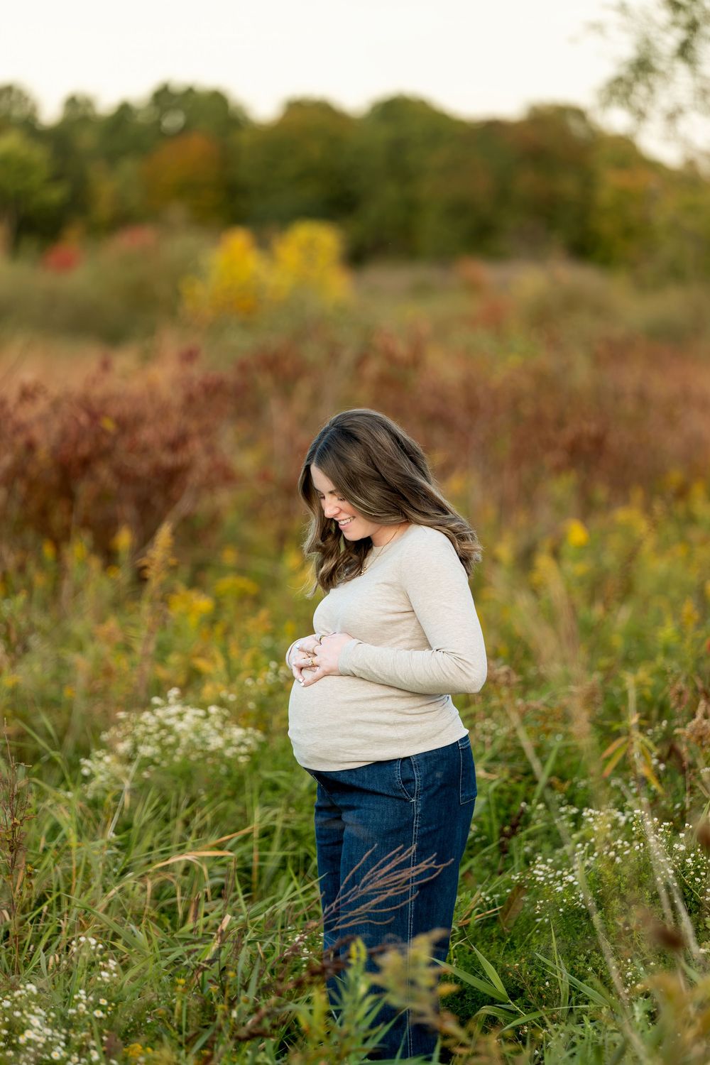 Pregnant woman in beige top and jeans cradles her baby bump while standing in a fall wildflower field.