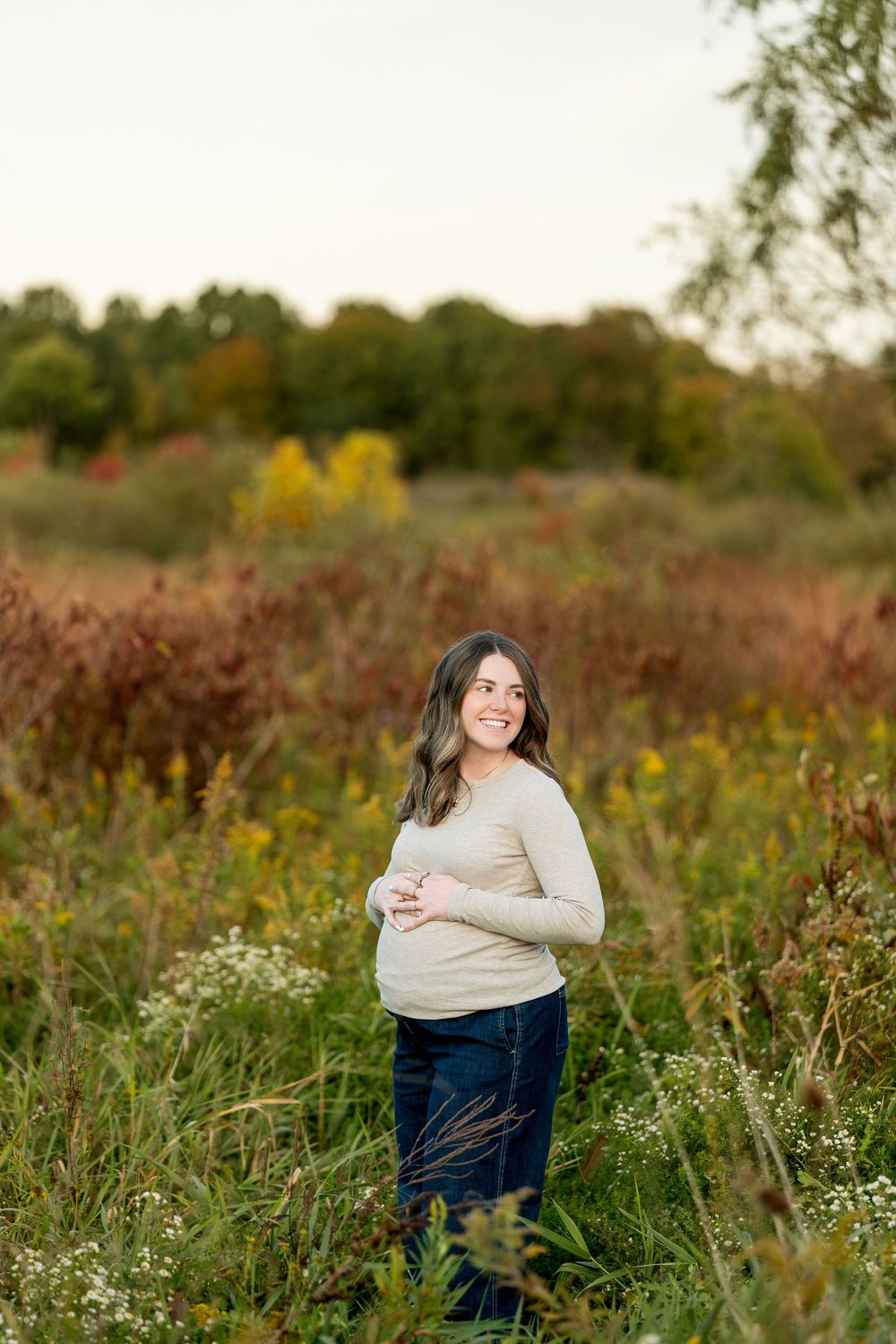 Smiling pregnant woman in beige top and jeans standing in a colorful autumn field during golden hour.