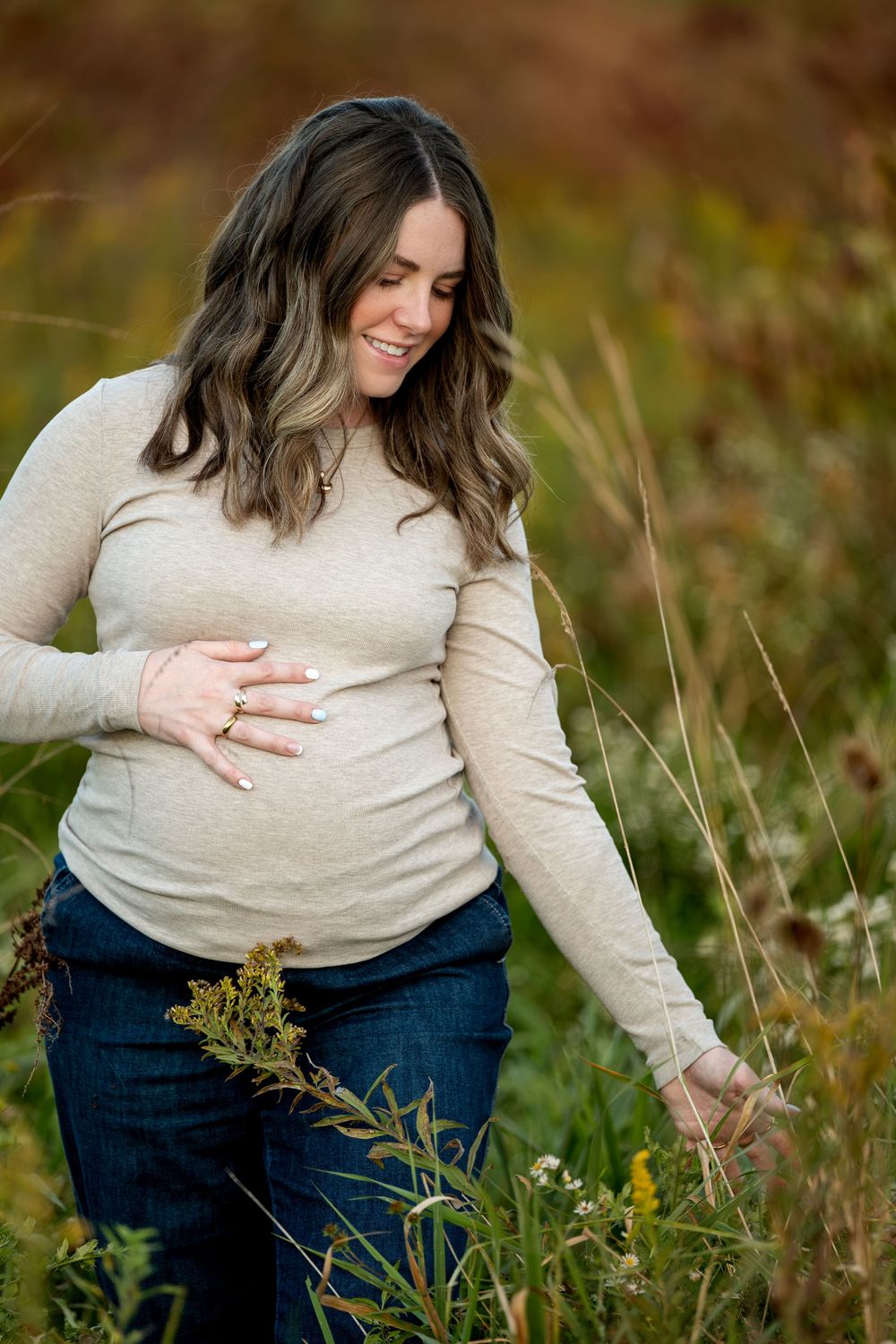 Smiling pregnant woman in beige top and jeans cradling her baby bump while touching wildflowers in an autumn field.