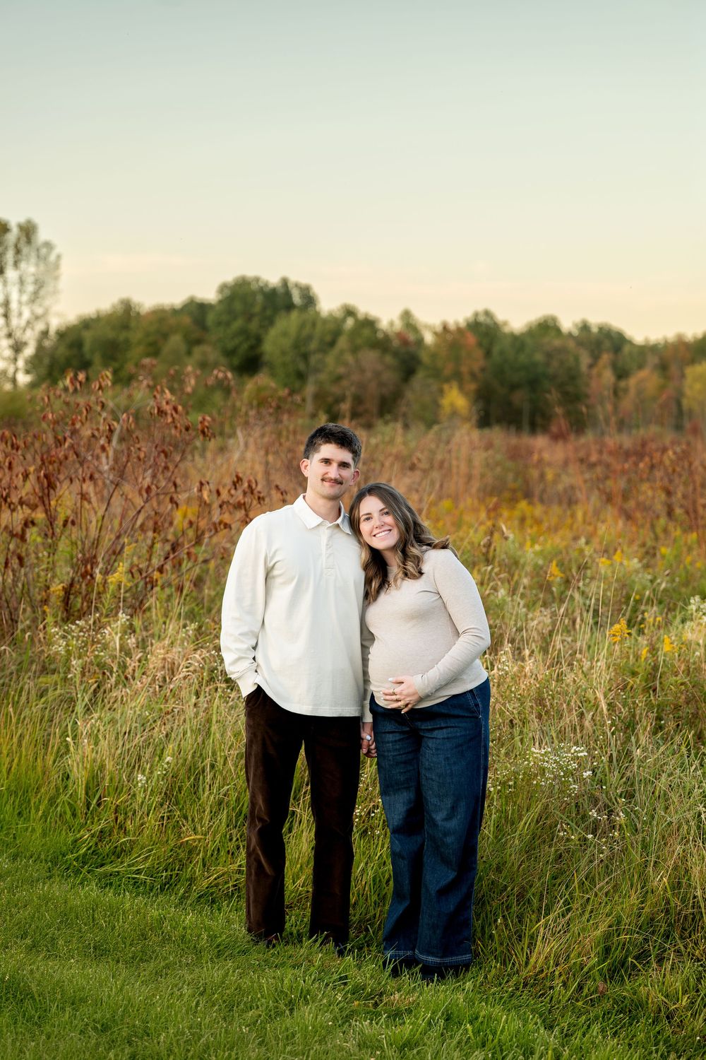 A couple poses in a field during golden hour, the woman cradling her baby bump in jeans and a beige sweater.