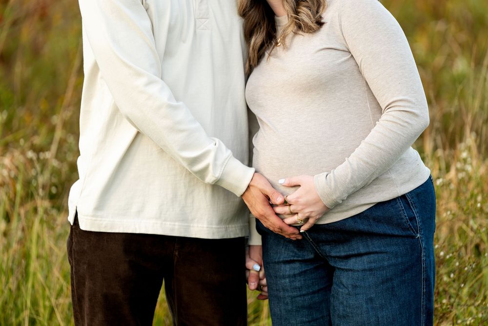 Couple tenderly holding pregnant belly together in a field, wearing casual neutral-toned clothing.