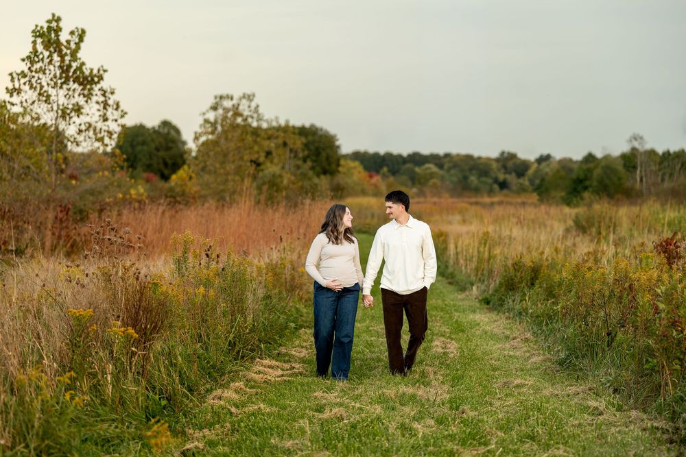 Couple walks hand in hand along a grassy path through a colorful autumn field during an outdoor engagement shoot.