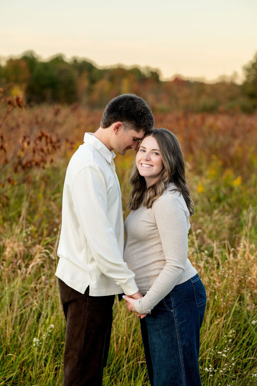 Couple holding hands smiling in autumn field at golden hour, woman in beige top and jeans, man in white shirt.