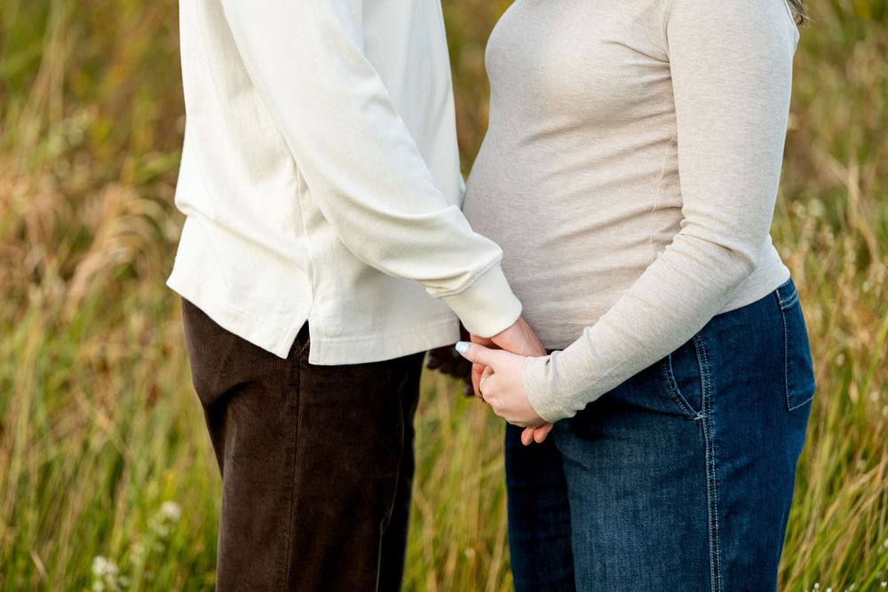 Pregnant couple holding hands in a field, woman showing baby bump in jeans and gray top.