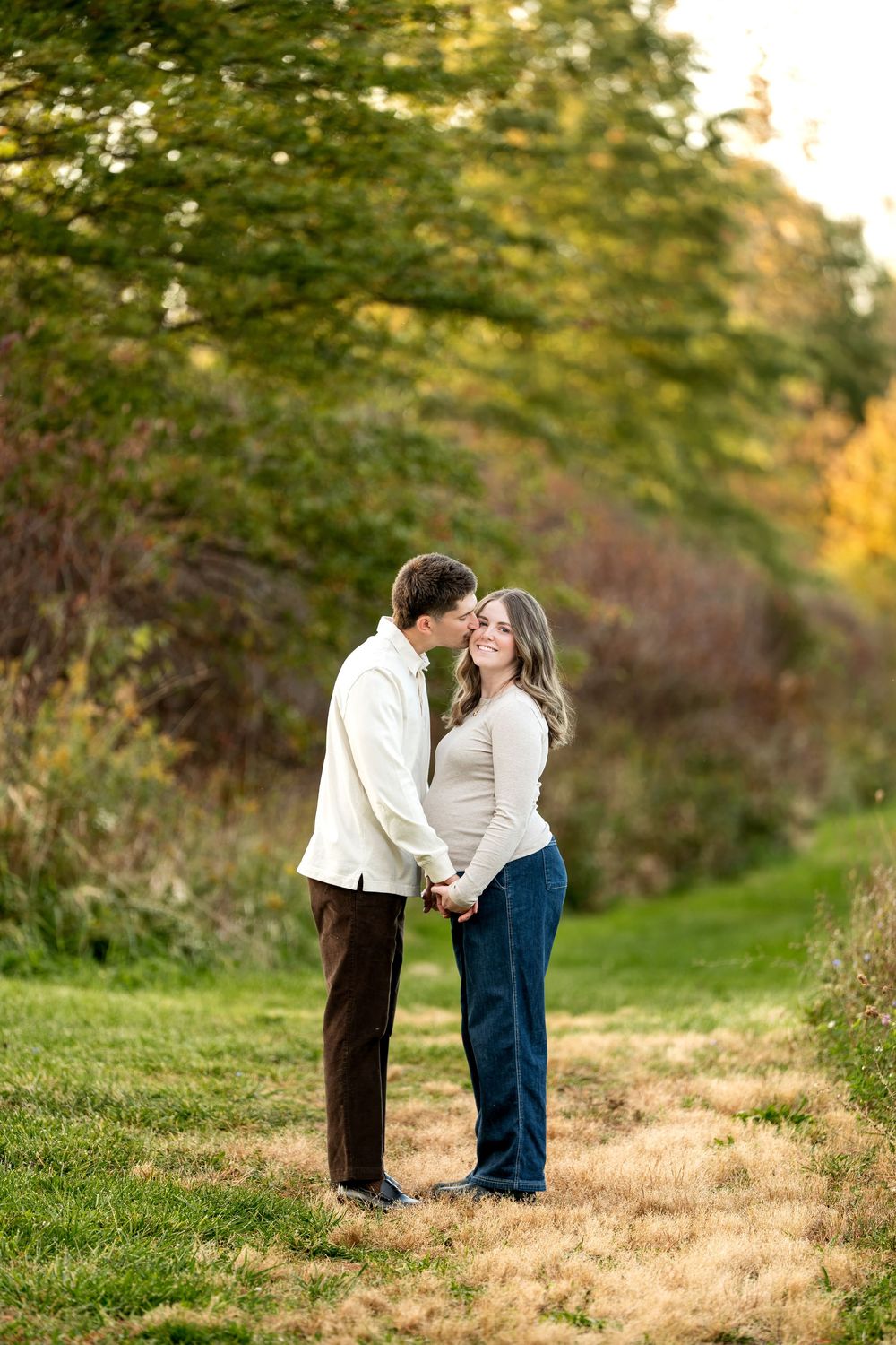 Couple holds hands and shares a kiss outdoors on a grassy path surrounded by colorful autumn foliage.